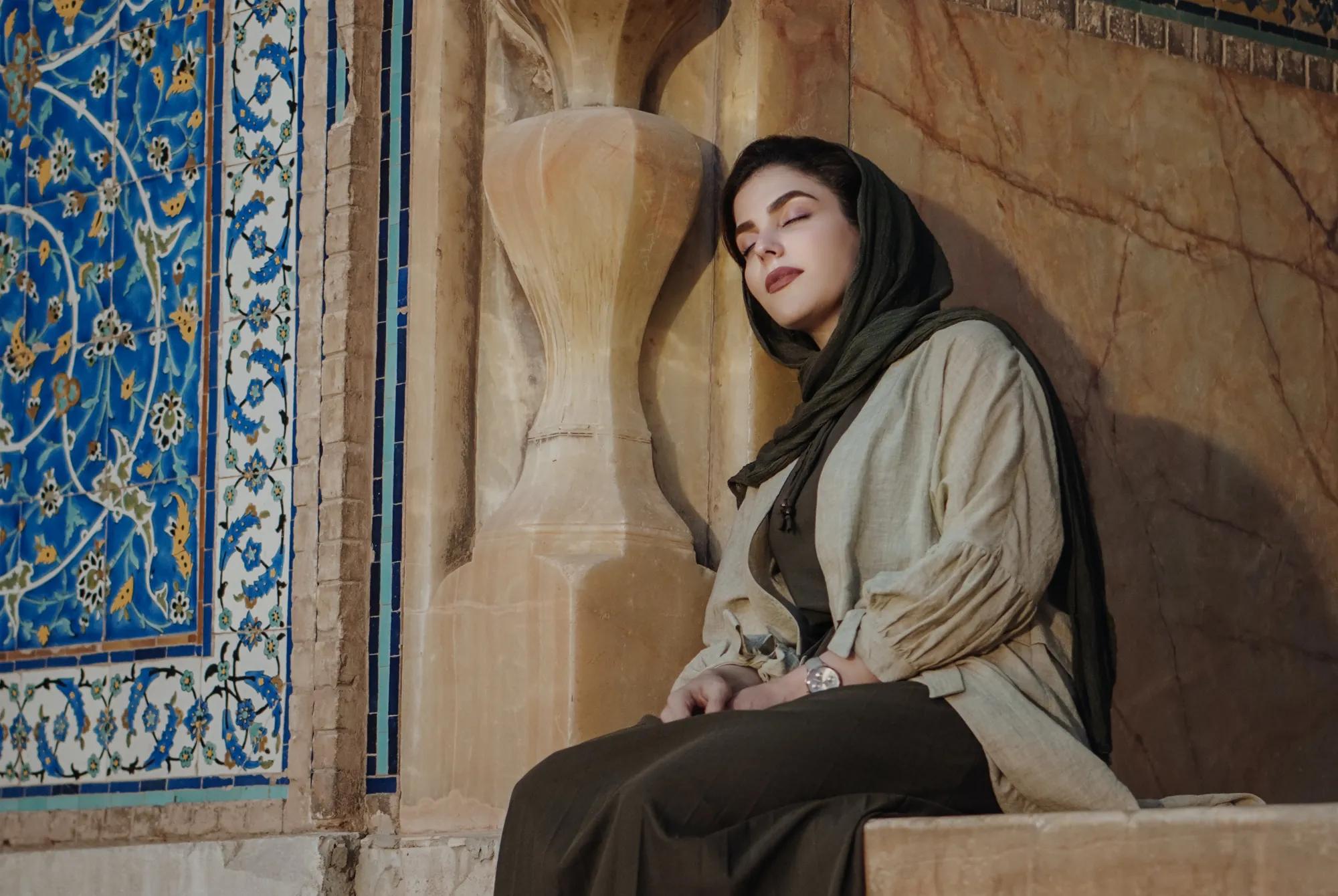 An iranian woman calmly resting against a wall with art